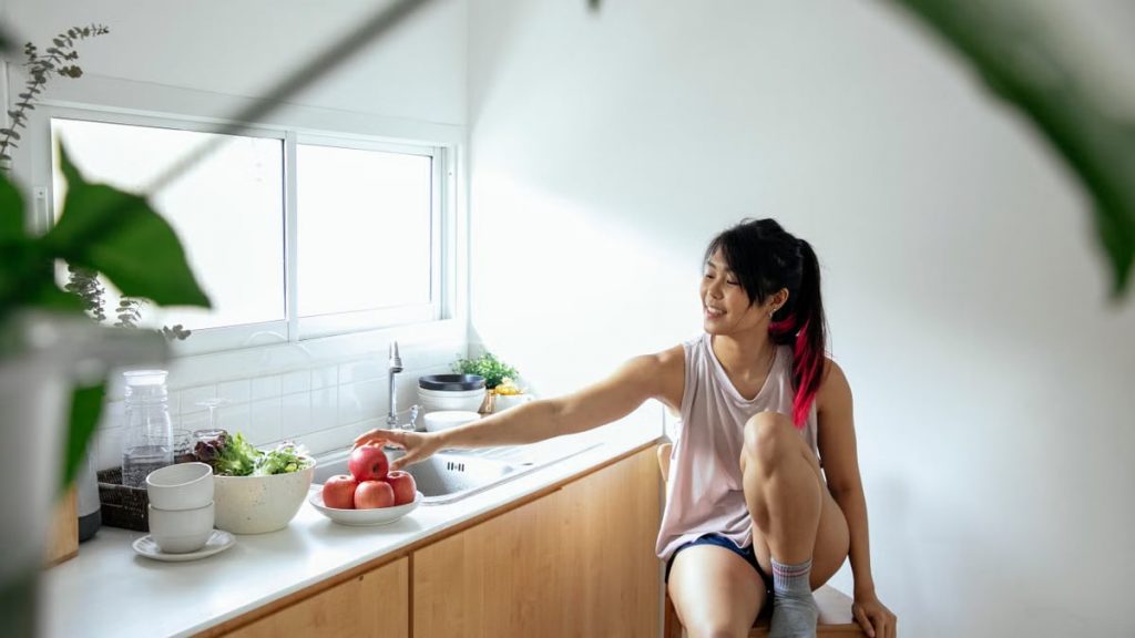Positive ethnic young lady in pajama sitting near counter with utensil and apples in plate near window in kitchen in daytime