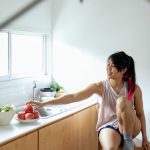 Positive ethnic young lady in pajama sitting near counter with utensil and apples in plate near window in kitchen in daytime