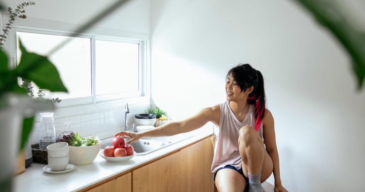 Positive ethnic young lady in pajama sitting near counter with utensil and apples in plate near window in kitchen in daytime